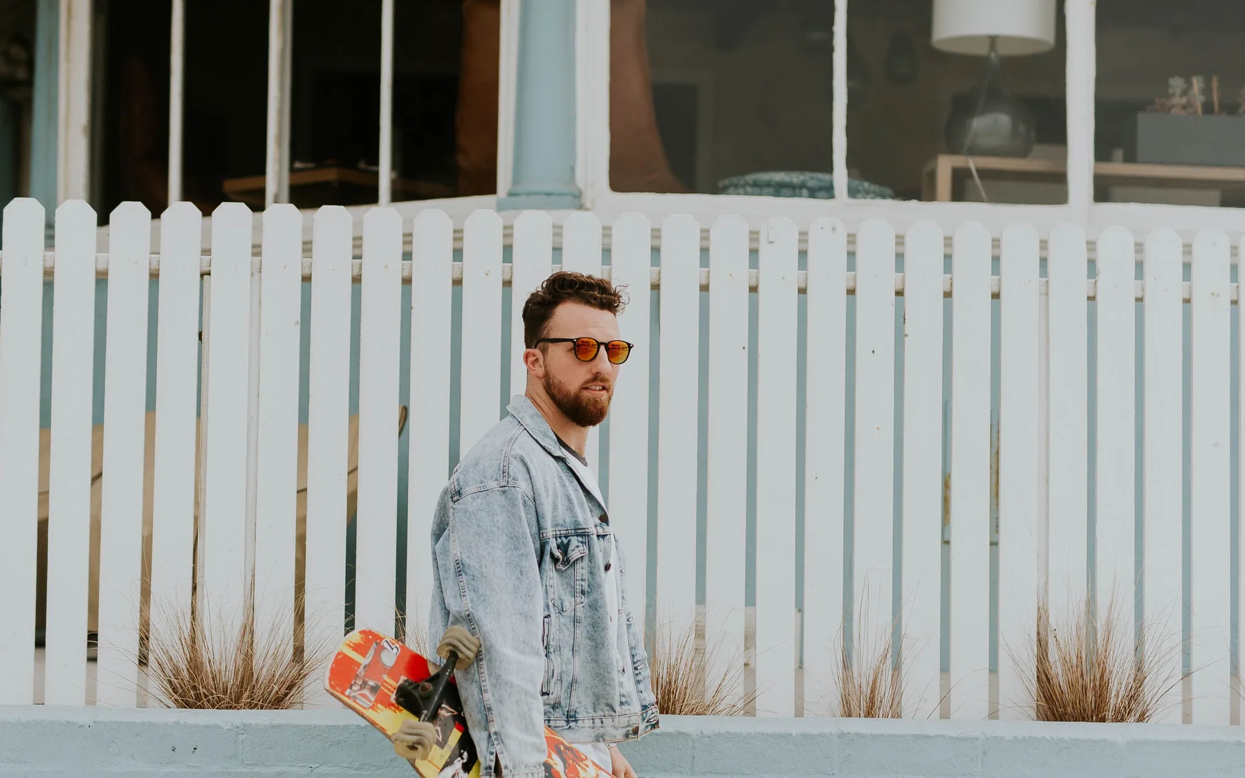 A man carrying a skateboard past a white fence